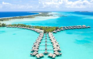 Overwater bungalows of The St. Regis Bora Bora Resort from overhead with crystal clear water in the lagoon and darker Pacific water outside.